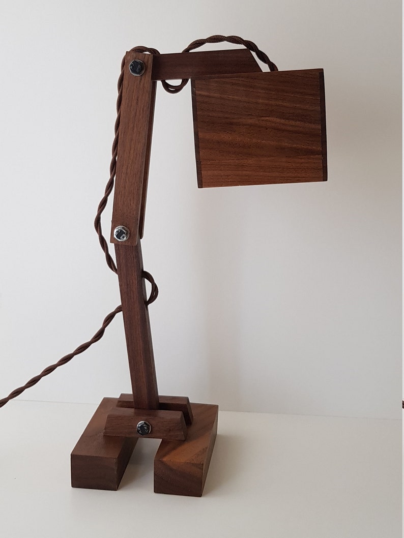 K&ouml;nnte beinhalten: Eine Schreibtischlampe aus Holz mit brauner Oberfl&auml;che. Die Lampe hat einen langen, beweglichen Arm und einen quadratischen Schirm. Die Lampe ist aus massivem Holz gefertigt und hat einen Vintage-Industriestyle.