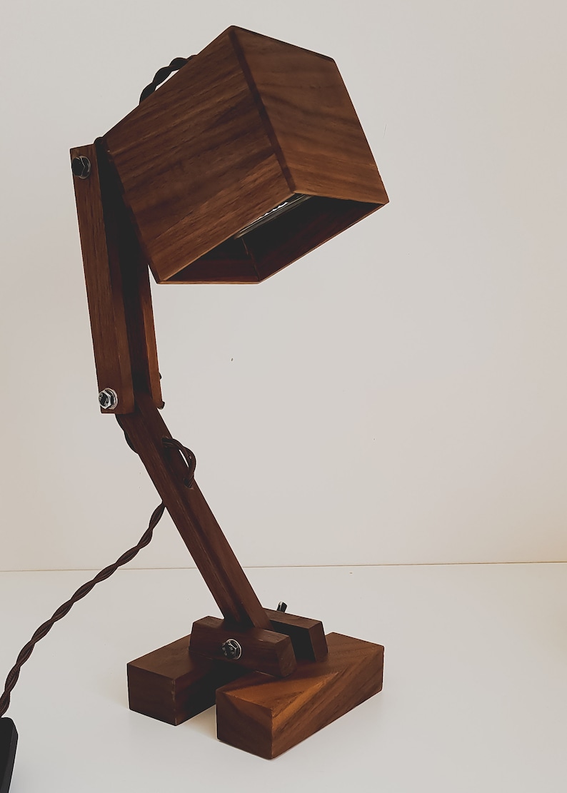 K&ouml;nnte beinhalten: Eine Schreibtischlampe aus Holz mit einem quadratischen Schirm und einem Gelenkarm. Die Lampe ist aus dunkelbraunem Holz gefertigt und hat ein schwarzes Kabel.