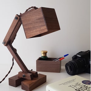K&ouml;nnte beinhalten: Eine Schreibtischlampe aus Holz mit einem quadratischen Schirm und einem verstellbaren Arm. Die Lampe ist aus dunkelbraunem Holz gefertigt und hat ein schwarzes Kabel. Die Lampe steht auf einem Tisch mit einem Buch, einer Kamera und einer kleinen Holzkiste.
