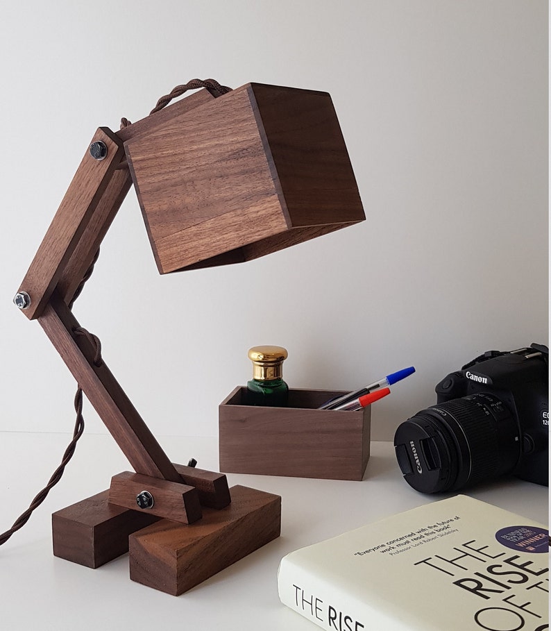 K&ouml;nnte beinhalten: Eine Schreibtischlampe aus Holz mit einem quadratischen Schirm und einem verstellbaren Arm. Die Lampe ist aus dunkelbraunem Holz gefertigt und hat ein schwarzes Kabel. Die Lampe steht auf einem Tisch mit einem Buch, einer Kamera und einer kleinen Holzkiste.
