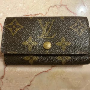 May include: A brown key holder featuring the iconic Louis Vuitton monogram pattern in a repeating gold-toned floral and LV logo design. The rectangular accessory includes a single gold-toned circular snap button closure centered on the front flap.