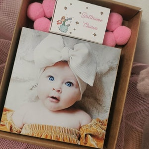 May include: A brown cardboard box containing a white card that reads "Battesimo Chiara" and a small photo of a baby girl wearing a white headband with a bow. The box also contains pink pom-poms.