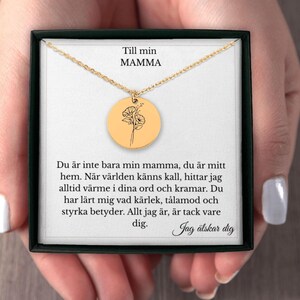 May include: A gold-colored necklace with a round pendant featuring a floral design. The pendant hangs from a delicate chain, presented in a black gift box. The text "Till min MAMMA" is visible.