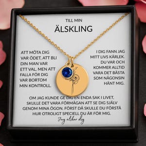 May include: A gold-colored necklace with a round pendant and a blue gemstone. The pendant has a floral design and text in Swedish. The necklace is presented in a black box with a white interior.