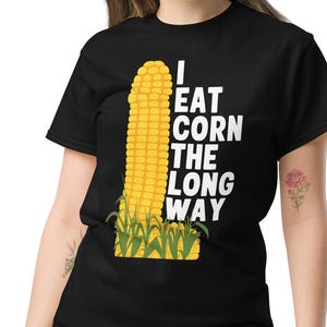 Op de afbeelding: Een zwart T-shirt met een witte afbeelding van een maïskolf en de tekst "I eat corn the long way".