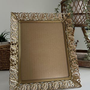 May include: A rectangular gold-coloured picture frame with an ornate, filigree design. The frame has a detailed floral and leaf pattern around the edges. The frame is empty, with a brown backing visible inside.