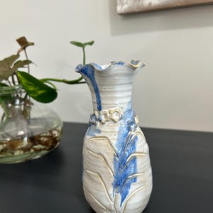 May include: A handmade ceramic vase with a ruffled rim and a textured surface. The vase features a cream-coloured base with blue accents and leaf-like patterns. The vase is approximately 15 cm tall.