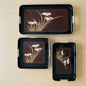May include: A set of three black rectangular and square serving trays with rounded corners and light yellow textured handles. Each tray features a central dark brown panel with a white and red floral design and a fine gold dot pattern background.