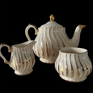 May include: A cream-coloured tea set comprising a teapot, a creamer jug, and a sugar bowl. Each piece features vertical gold stripes and gold detailing. The teapot has a gold finial on its lid. The set is designed for serving tea.