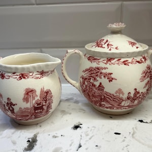 Peut inclure: Ensemble de pot à crème et sucrier couverts en céramique crème. Les deux pièces présentent un motif rouge de paysage avec des personnages et des bâtiments. Le sucrier a un couvercle avec un bouton floral et des poignées.