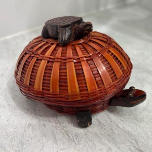 May include: A turtle-shaped basket, featuring a woven reddish-brown body and dark wooden turtle head and feet. The lid is a small wooden turtle. This decorative basket is likely for storage or display.