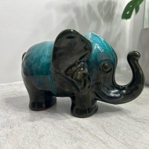 May include: A ceramic elephant ornament with a teal and black glaze. The elephant has a curved trunk, large ears, and a detailed texture. The figurine is a decorative item, suitable for home decor.