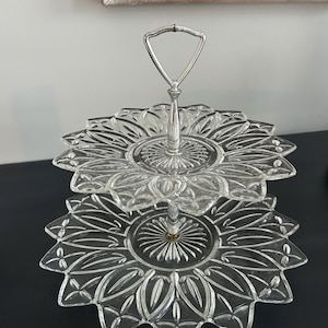 May include: A two-tiered, clear glass serving tray with a silver handle. The trays are shaped like flowers with pointed petals. The serving tray is ideal for displaying desserts or appetizers.