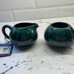 May include: A matching set of ceramic cream and sugar bowls. The cream jug has a handle and spout, while the sugar bowl is round. Both are glazed in a dark green colour with a glossy finish and a dark interior. A tag is attached to the cream jug.
