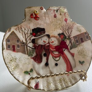 May include: A shell-shaped decorative dish depicting a winter scene with two snowmen wearing scarves and hats. The dish has a gold-coloured rim and is supported by a gold-toned metal stand. The background shows a snowy landscape with houses and trees.