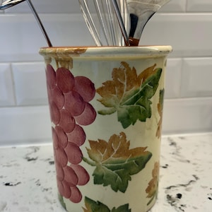 May include: A cream-coloured ceramic utensil holder decorated with a bunch of red grapes and green and brown leaves. The holder contains stainless steel kitchen tools, including a whisk and spatula.