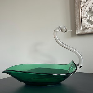 Peut inclure: Un bol décoratif en forme de cygne en verre. Le bol est vert émeraude foncé avec un bord noir, et le cou et la tête du cygne sont en verre transparent. Le bol est posé sur une surface sombre.