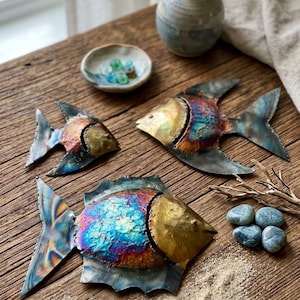 May include: Three metal fish sculptures with iridescent blue, gold, and red hues are displayed on a wooden surface. The fish have detailed fins and scales. A small ceramic bowl with marbles, a vase with greenery, and pebbles are also present.