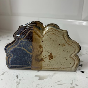Peut inclure: Un porte-serviettes en céramique avec un design festonné unique. Le support est divisé en deux sections, l'une bleu foncé et l'autre beige moucheté. La pièce a une finition brillante et une esthétique rustique et artisanale.