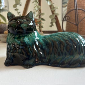 May include: A ceramic cat figurine in a reclining position, featuring a glossy glaze with blended shades of deep forest green, teal, and black. The surface has a textured, wavy pattern across the body, with smooth, rounded ears and facial features.
