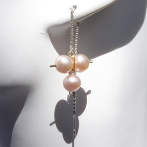 Puede incluir: Un solo pendiente con tres perlas de color rosa pálido suspendidas de una fina cadena de plata. El pendiente se muestra sobre un fondo blanco, con la sombra del pendiente visible.
