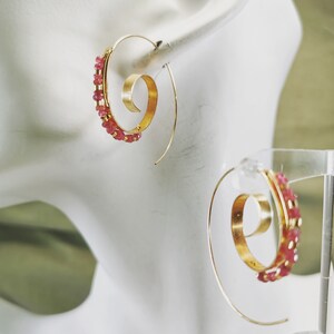May include: Gold-tone spiral earrings with a row of small, pink gemstones. The earrings have a unique, open design with a curved hook for the ear. The gemstones are a vibrant pink color, adding a pop of color to the gold metal.