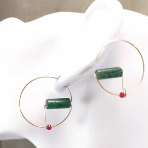 May include: Gold-coloured hoop earrings with a geometric design. Each earring features a green cylindrical stone and a small red bead. The earrings are displayed on a white surface.