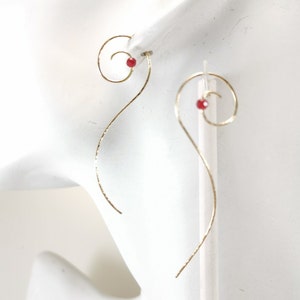 May include: A pair of gold-colored earrings with a spiral design. Each earring features a small, round, red bead at the top of the spiral. The earrings have a long, curved, hammered texture. The earrings are displayed on a white surface.