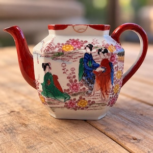 Puede incluir: Una tetera de cerámica blanca con cuerpo hexagonal, que presenta un asa y un pico de color rojo. La superficie está decorada con una colorida ilustración de tres figuras con vestimenta tradicional entre motivos florales rosas y amarillos.