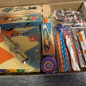 May include: A box filled with vintage toys and candy. Items include a wooden train whistle, a comb, a metal kazoo, a harmonica, a yo-yo, lollipops, and wrapped candies. The packaging features retro designs with bright colors and bold text.