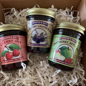 May include: Three jars of Cooper's Mill jam in a cardboard box with white paper filler. The jars are labeled Cherry Jam, Blueberry Lavender Jam, and Cranberry Apple Jam. Each jar has a gold lid and a label with fruit illustrations.