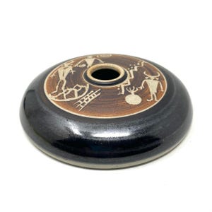May include: A black ceramic vase with a brown rim and white painted designs of Native American figures and symbols. The vase has a round shape and a small opening at the top.