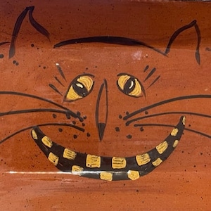 Peut inclure: Un plat en céramique terre cuite avec un motif peint à la main d'un visage de chat. Le chat a des moustaches noires, des yeux jaunes et un large sourire aux dents noires et jaunes.