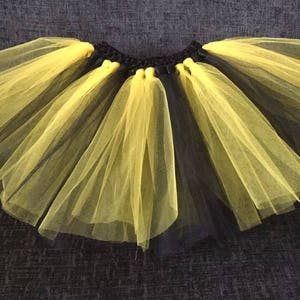 Può includere: Una gonna tutu in tulle nero e giallo. La gonna è realizzata con più strati di tulle e ha una fascia elastica nera.