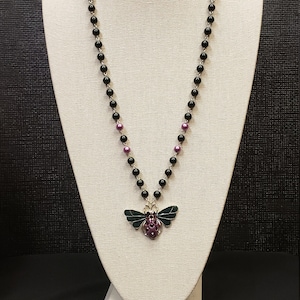 Op de afbeelding: Een zwarte en paarse kralenketting met een groene en paarse bij-hanger.
