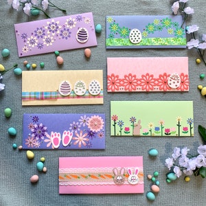 May include: A selection of Easter-themed greeting card envelopes in various colours. Each envelope is decorated with floral patterns, Easter eggs, and bunny motifs. The envelopes are arranged on a blue surface, with additional Easter decorations.
