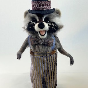 May include: A needle-felted raccoon figurine wearing a black top hat with a decorative purple band and a white feather. The raccoon has a floral print bow tie, light blue suspenders, and brown and purple striped trousers with two buttons.