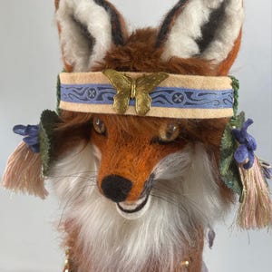 May include: A handcrafted fox figurine with reddish-brown and white fur, wearing a beige headband featuring a blue patterned ribbon and a gold-colored butterfly emblem. The fox has dark eyes, a black nose, and decorative tassels with purple fabric leaves.