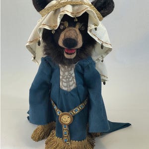 May include: A needle-felted bear figurine wearing a cream-colored head covering with gold trim and a blue corduroy robe. The robe features a gold-toned belt with a circular buckle, a light-colored patterned chest panel, and a gold fringe hem.