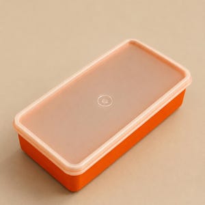 Puede incluir: Un recipiente de almacenamiento rectangular con una base de color naranja vibrante y una tapa semitransparente de color melocotón esmerilado. La tapa presenta una pequeña hendidura circular centrada que asegura el cierre de la caja naranja.