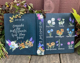 Puede incluir: Un diario negro con una cita de Lucas 12:27, "Considera las flores silvestres, cómo crecen." La portada está decorada con flores silvestres pintadas y sus nombres, incluyendo margaritas de ojo de buey, cardos, lavanda, corendon, caléndulas, nomeolvides, rudbeckias, campanillas y cosmos.