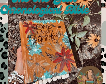 Puede incluir: Un libro encuadernado en cuero marrón con un diseño floral en la portada. El título "The One Year Chronological Bible" está escrito en letra negra en la portada. El libro está sostenido en una mano con una pulsera en la muñeca.