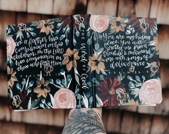 Puede incluir: Un libro negro con un diseño floral pintado en la portada. El libro está abierto para revelar las palabras "The Word of God" escritas en el lomo. Las páginas están llenas de texto escrito a mano y versículos bíblicos, incluyendo "Salmo 103:13" y "Salmo 32:7".