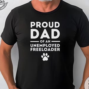 May include: Black t-shirt with white text that reads "PROUD DAD of an UNEMPLOYED FREELOADER" and a paw print. The shirt has the words "AUTHENTIC COMFORT COLORS 100% COTTON" and "SHIPS FAST!" printed on it.