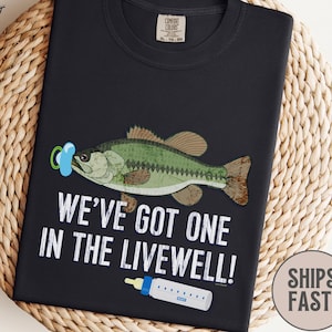 May include: Black t-shirt with a graphic of a fish wearing a pacifier and a baby bottle. The text reads "WE'VE GOT ONE IN THE LIVEWELL!". The shirt is made of 100% cotton.