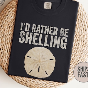 May include: Black t-shirt with the text "I'D RATHER BE SHELLING" and a sand dollar graphic. The shirt is made of 100% cotton and has a Comfort Colors label. The shirt is displayed on a woven surface.