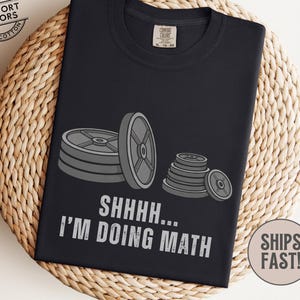 May include: Black t-shirt with a graphic of stacked weight plates in shades of gray. The text "SHHHH... I'M DOING MATH" is printed in white. The shirt is made of 100% cotton.