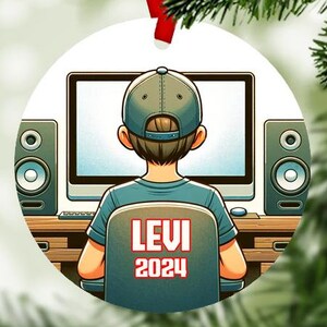 May include: A white round Christmas ornament with a cartoon illustration of a person sitting in front of a computer with headphones on. The person is wearing a black and red baseball cap and a blue shirt. The text "LEVI 2024" is on the back of the chair.