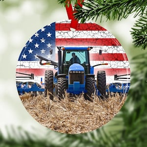 May include: A blue tractor with a red, white, and blue American flag background. The tractor is driving through a field of dry, brown grass. This is a Christmas ornament.