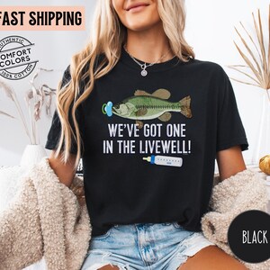 May include: Black t-shirt with the text "WE'VE GOT ONE IN THE LIVEWELL!" featuring a cartoon bass wearing a pacifier and a baby bottle. The shirt is made of 100% cotton.